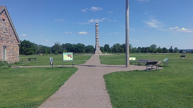 Path in a grassy field leading to a building at left, a flag pole at center right, and a pillar in the background.